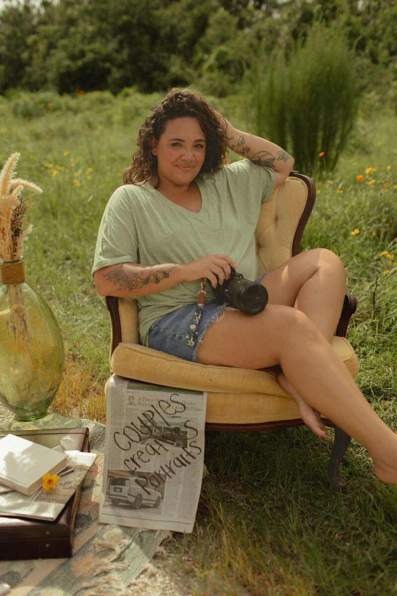 Florida couples and portrait photographer sitting in a vintage chair in a wildflower field, holding a camera with a newspaper labeled "couples, creatives, portraits" beside her.