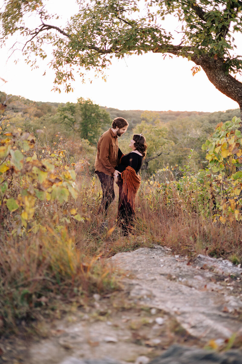 Expecting couple standing in a field with a tree at sunset looking at each other lovingly. 