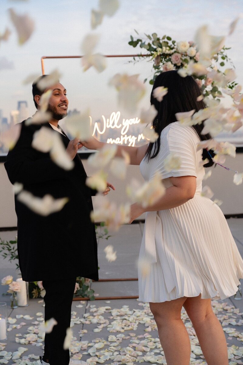 Couple surrounded by falling rose petals with a glowing 'Will you marry me?' sign in the background, styled by Yours Truly, a wedding planner and designer in New Jersey.