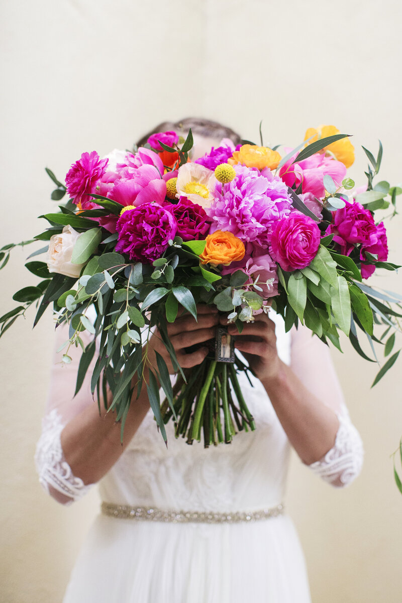 A bride holds a vibrant bouquet of pink, orange, and lush green florals, showcasing beautiful wedding flower arrangements by WINK! Weddings.