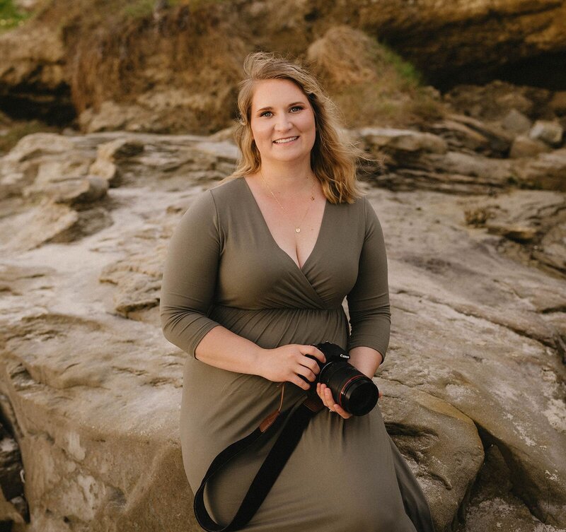 Molly, a San Diego photographer, sitting on rocks with her camera in hand, smiling at the camera