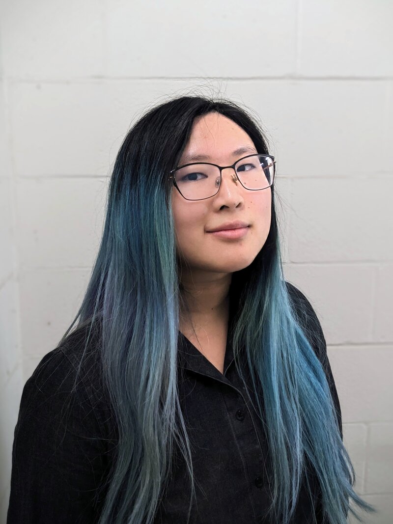 A woman with long, straight hair dyed in shades of blue and black, wearing glasses and a black button-up shirt. She is standing against a light-colored cinder block wall, looking directly at the camera with a neutral and confident expression.