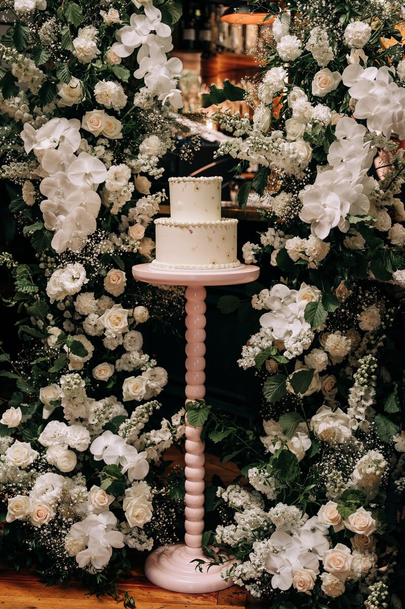 a cake with pressed flowers by fulltime tart on pink cake stand with white floral pillars around it by the flower press nz at 5th street restaurant for wedding reception