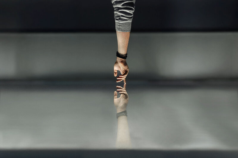 The image shows a pair of legs raised vertically in the air, wearing light blue jeans and black Converse-style sneakers with white laces and soles. The legs emerge from dense green foliage or plants in the foreground. The background is blurred, showing an out-of-focus wooded area with tall, leafless trees. The scene has a natural, outdoor setting.