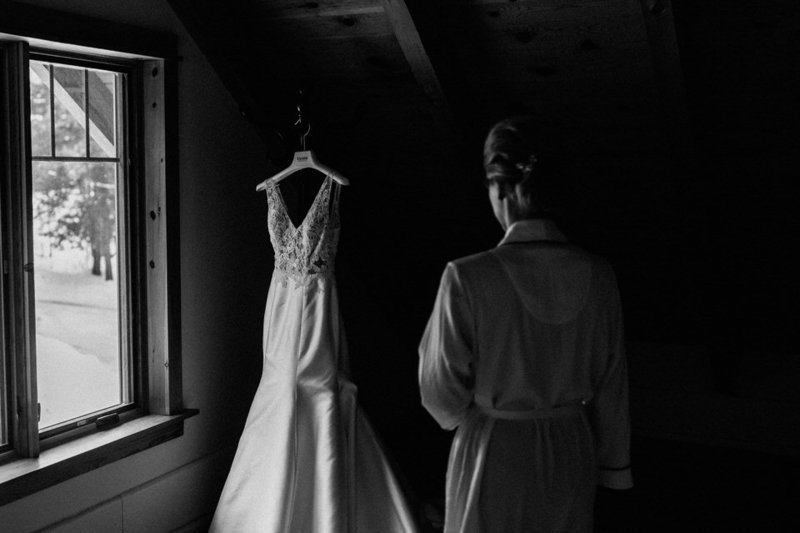 A black and white photo of a bride looking at her wedding dress hanging on rustic wooden beams,