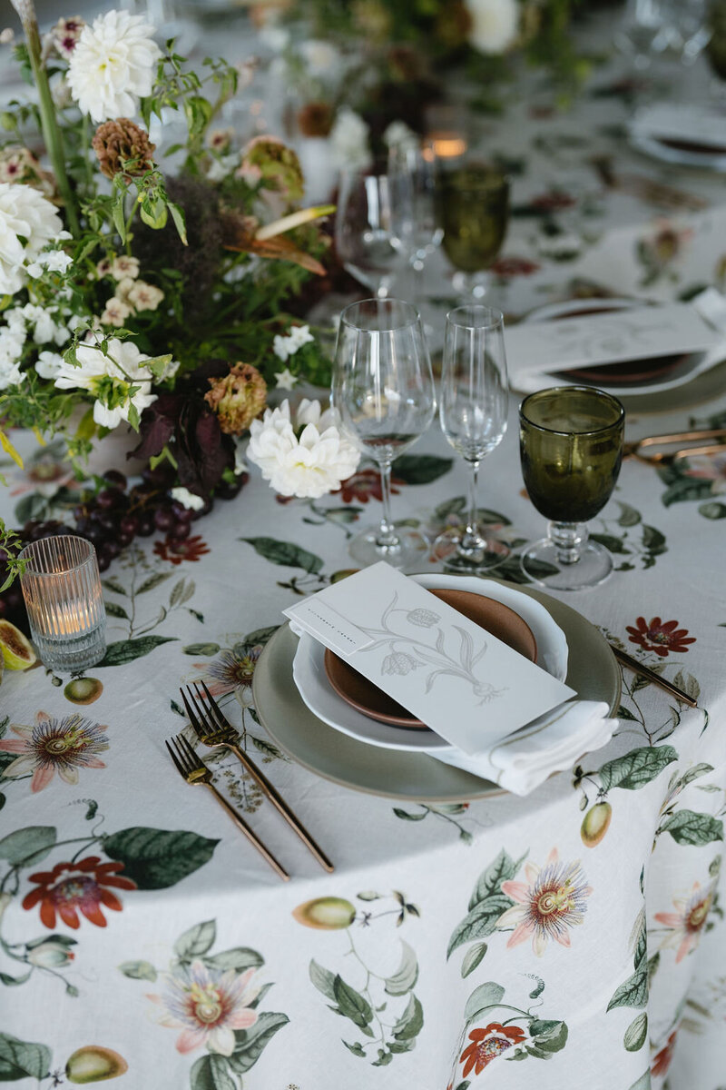A beautifully set table featuring botanical-patterned linens, floral centerpieces, gold flatware, and green glassware.