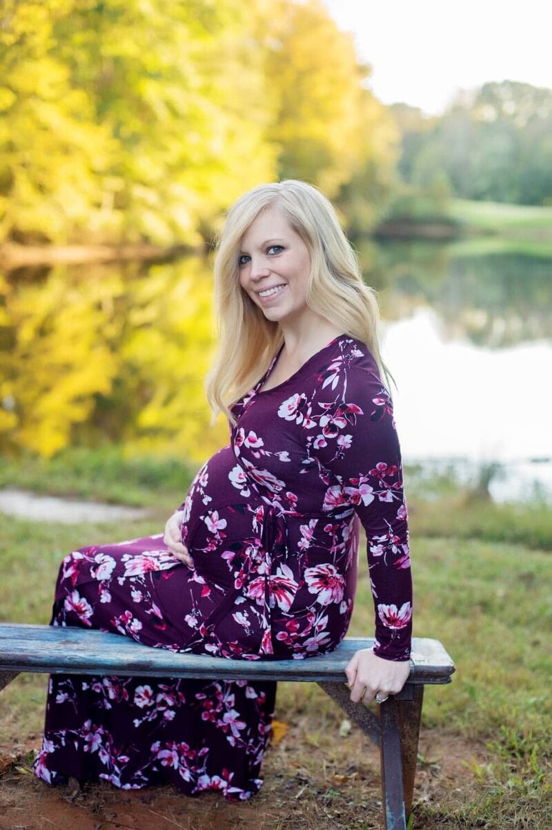 Happy mother-to-be in a purple and pink floral dress sitting on the park bench in front of a pond.