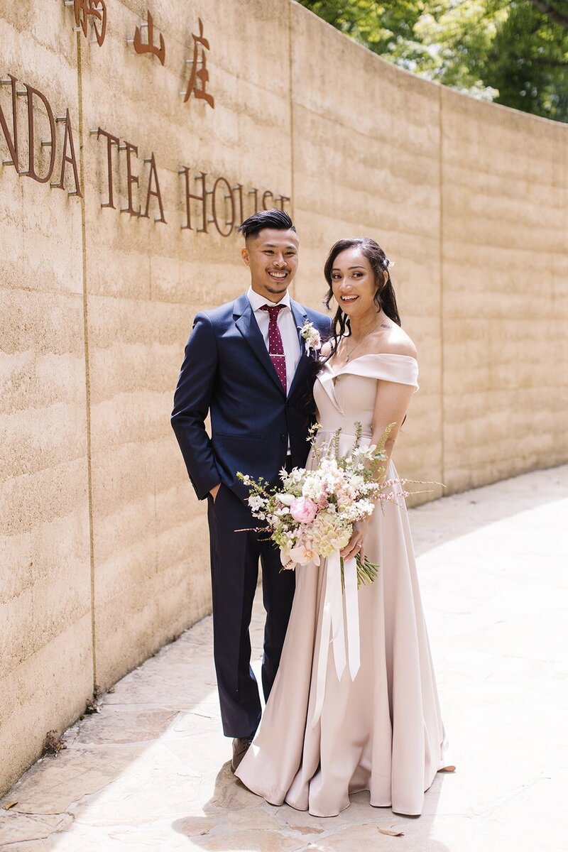 A bride in a champagne coloured dress and a groom in a blue suit standing in front of a curved wall that says Olinda Tea House