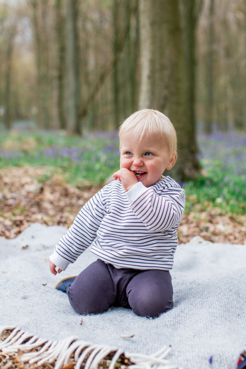 Little blonde boy sitting on a grey rug in a bluebell forest with a cheeky smile
