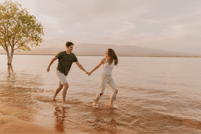 A man and a woman holding hands while running through water. The sane is orange and their is a tree behind them to the left.