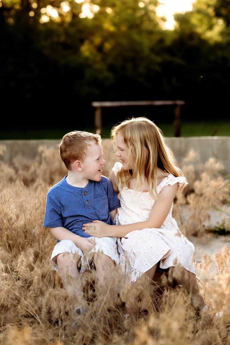 Two children, a boy and a girl, sitting on a box in the summer foliage laughing and smiling at each other.