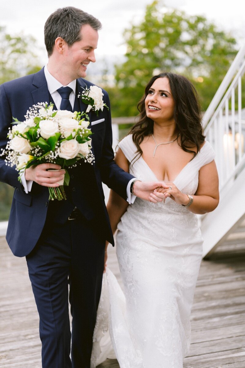 A bride and groom holding hands and smiling at each other on a bridge, the groom holding a bouquet of white flowers.