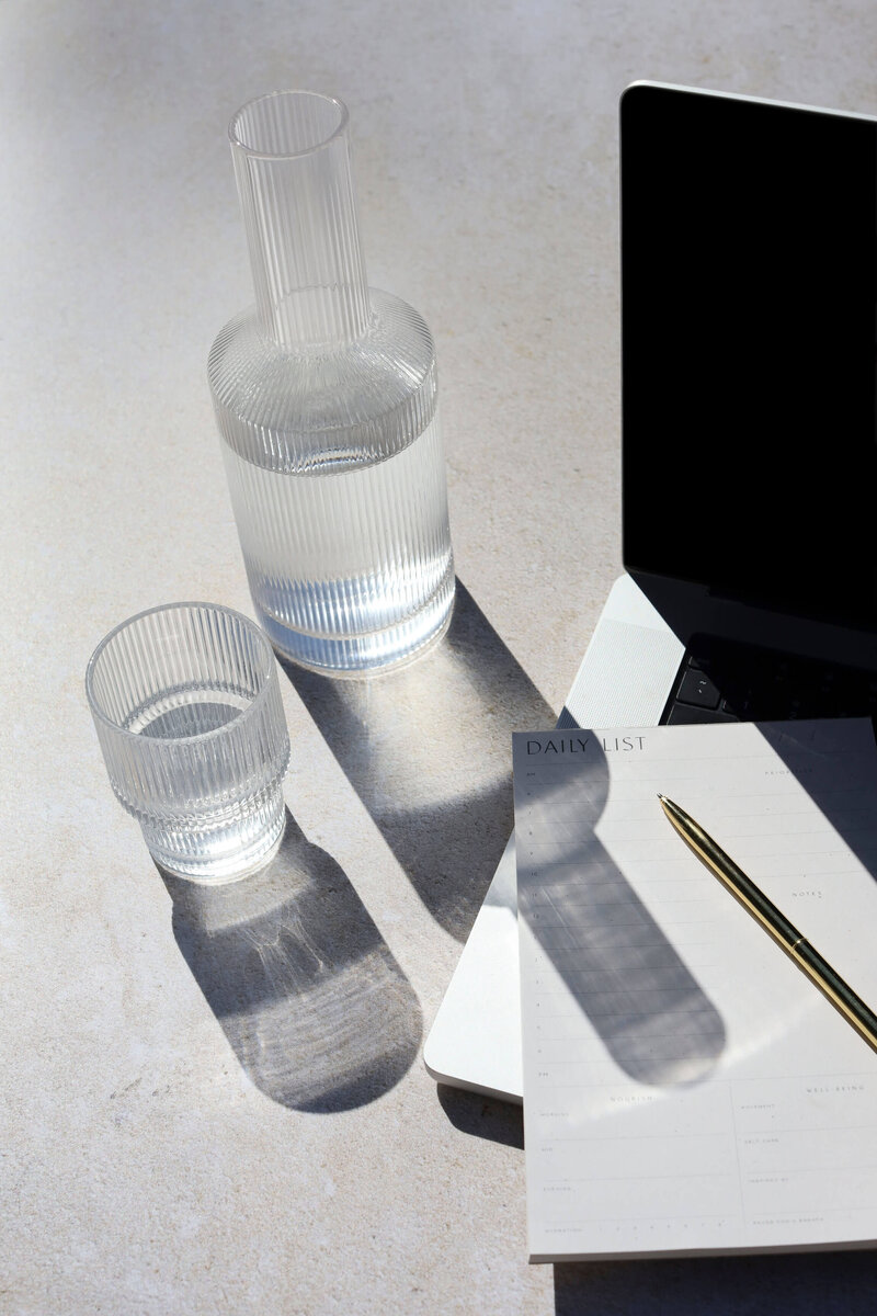 A laptop, a water jug, and a glass of water sit next to a writing pad.