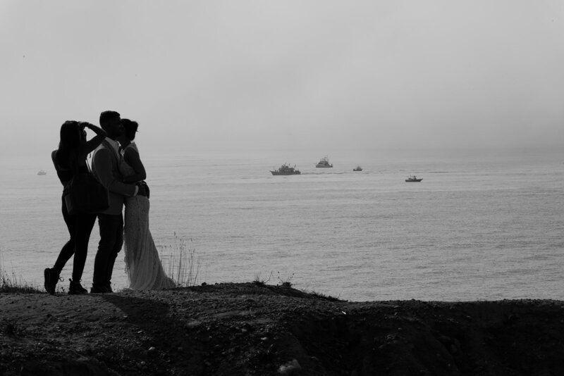 A stylish woman sits on a spiral staircase against a rustic brick wall, holding a film camera. She's dressed in a white blazer, black lace top, designer belt, and strappy black heels, exuding effortless confidence and creative energy.
