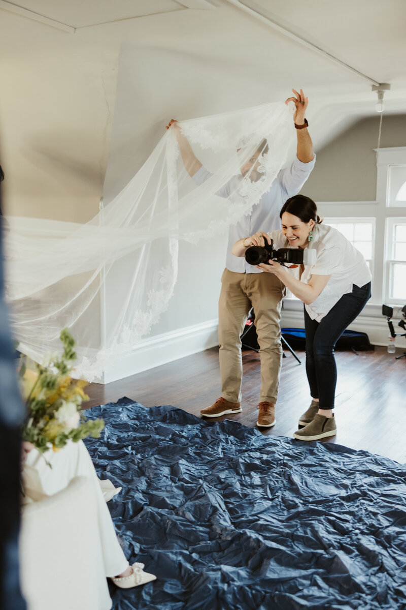 A photographer smiling as someone lifts up the end of a veil as they take a picture 