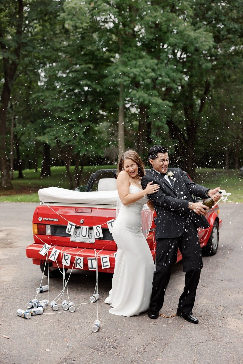 bride and groom on wedding day pop champagne bottle and squint to avoid foam standing next to red convertible volkswagon with just married sign and cans on back image by minnesota elopement photographer tom thornton photography