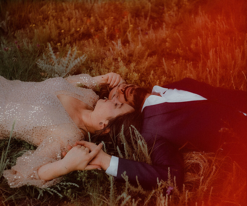 A bride and groom lay in a field holding hands with their heads next to each other
