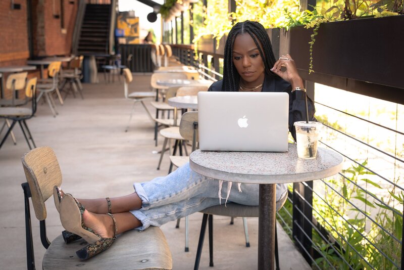 Rianne Nycole sitting at a table in a black shirt and jeans.  She is at Onyx coffee lab with her feet crossed and propped on a nearby chair. She is looking down at her laptop and has a plastic cup of water on the table.
