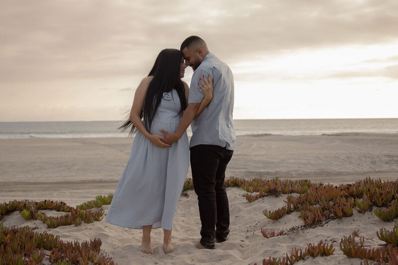 Expecting couple standing on the beach, forehead to forehead, with the mother holding her baby bump
