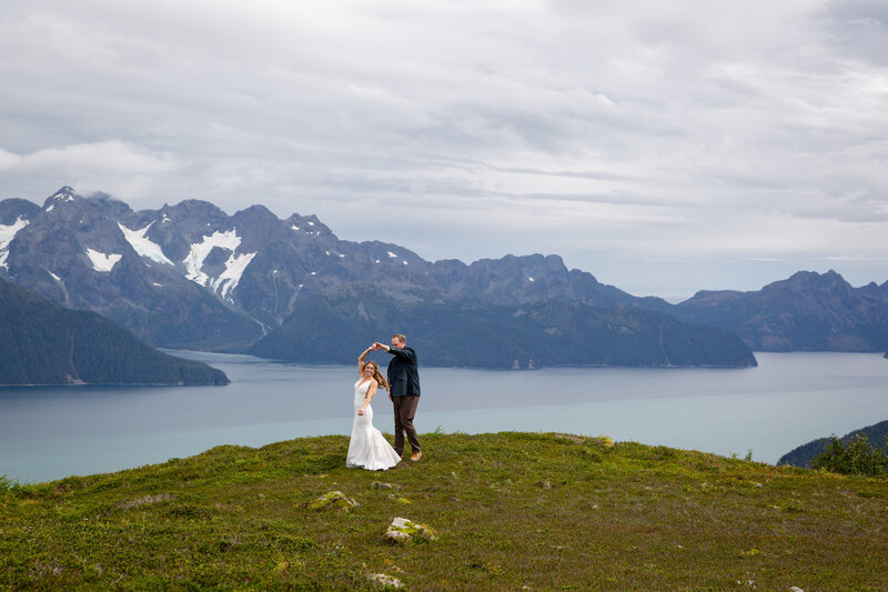A bride in a white dress spins under her grooms arm as he twirls her on top of a grassy hill in Alaska.