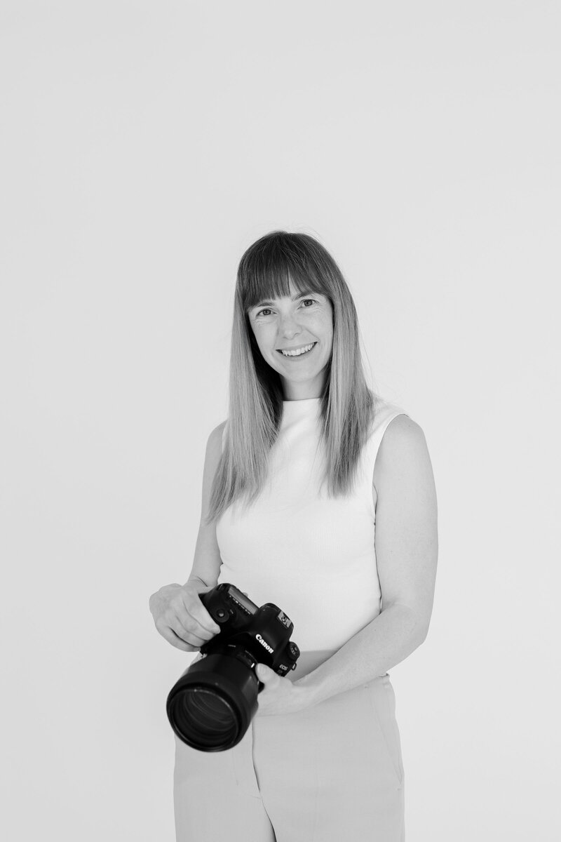 Black and white portrait of Katie Crane, a Toronto-based family photographer, smiling while holding a Canon camera. Known for capturing soulful, emotive, and timeless images, Katie specializes in newborn, lifestyle, and outdoor family photography in the Toronto and Durham regions. This image reflects her warm, storytelling approach to photography.