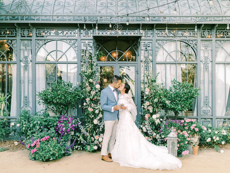 A couple in wedding attire shares a kiss in front of a vintage greenhouse adorned with lush greenery and colorful flowers.