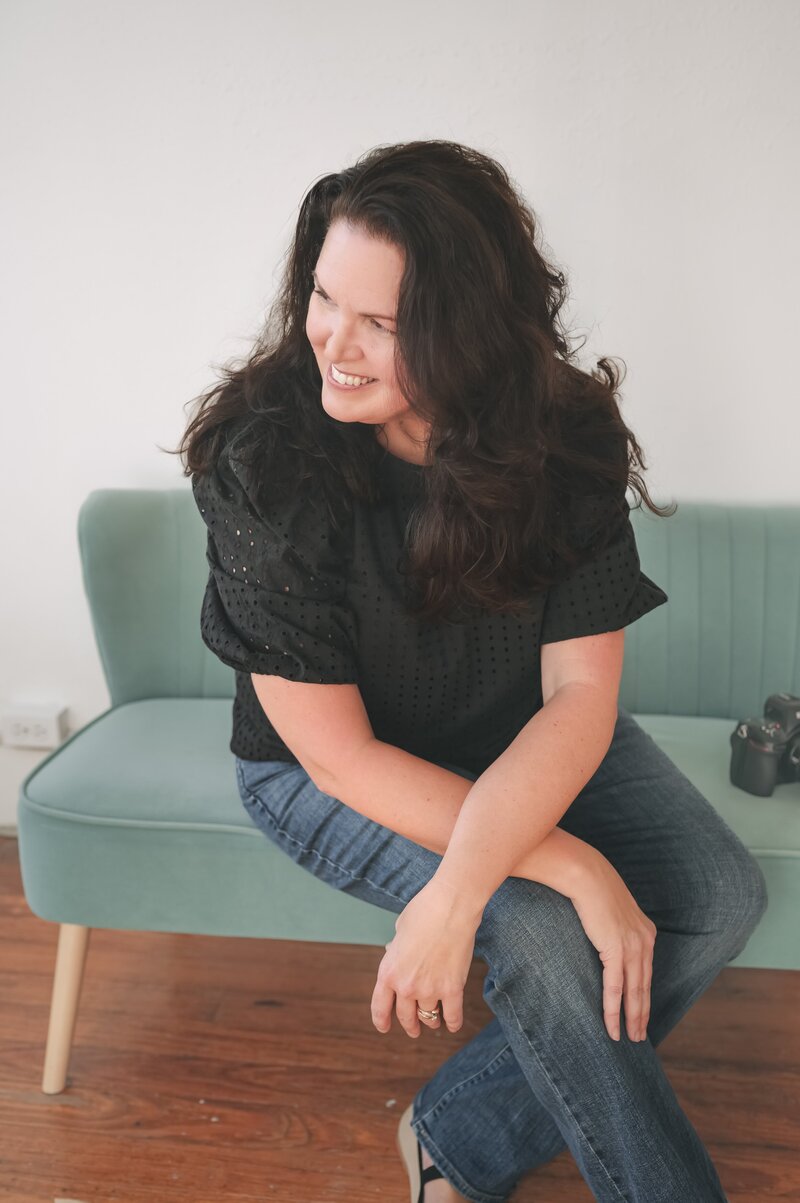 Jennifer sitting on couch with camera beside her, smiling and looking to the side during personal branding session in New Braunfels, Texas.