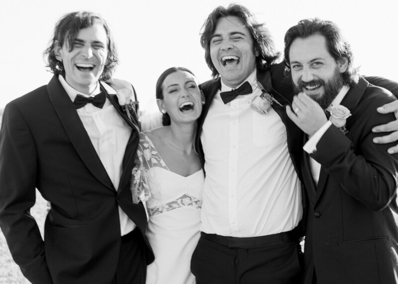 Black and white image of bride and groomsmen laughing with their arms around each other