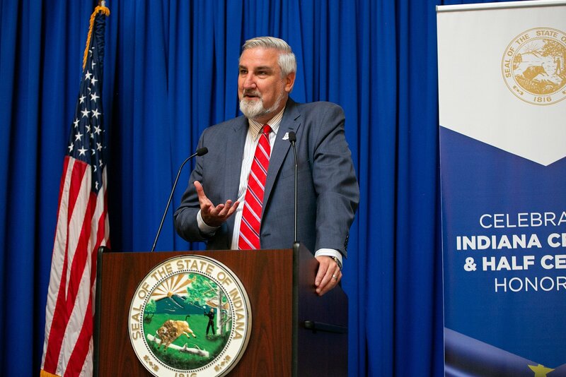 a-government-official-speaking-at-a-podium-with-the-indiana-state-seal-and-american-flag-during-a-press-conference-in-indianapolis-at-the-indiana-statehouse