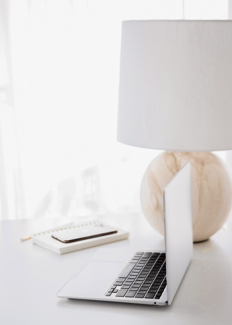 A laptop angled and open with a lamp and phone and notebook in the background, all placed on a white table