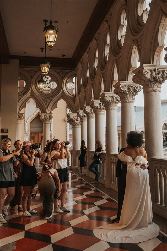 A group of photographers captures a moment between a couple embracing in an ornate hallway with tall, arched windows and columns in Las Vegas at The Venetian.