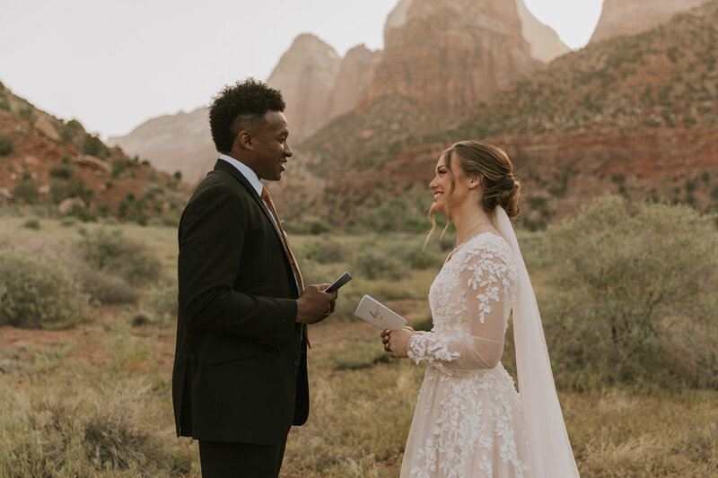 A bride and groom standing across from each other, smiling. Both are holding their vow books and have the landscape of Southern Utah behind them.