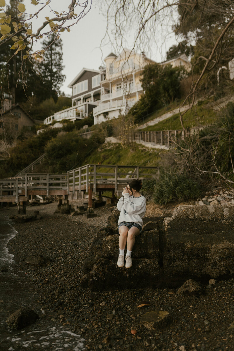 Woman sitting on rock by coastline taking photos of the ocean with coastal cliffs and houses in the background