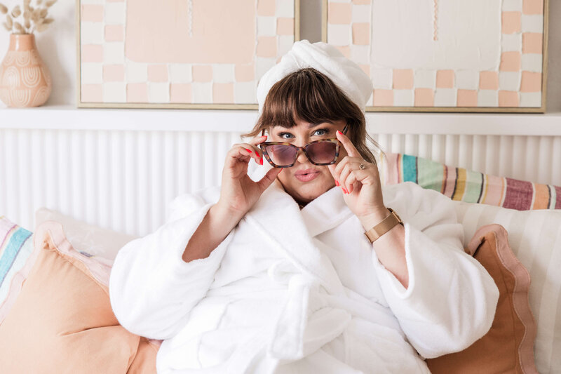 Leith in a terry towelling robe, with a towel on her head, wearing tortoiseshell glasses, holding them down the bridge of her nose and looking over them while pouting.