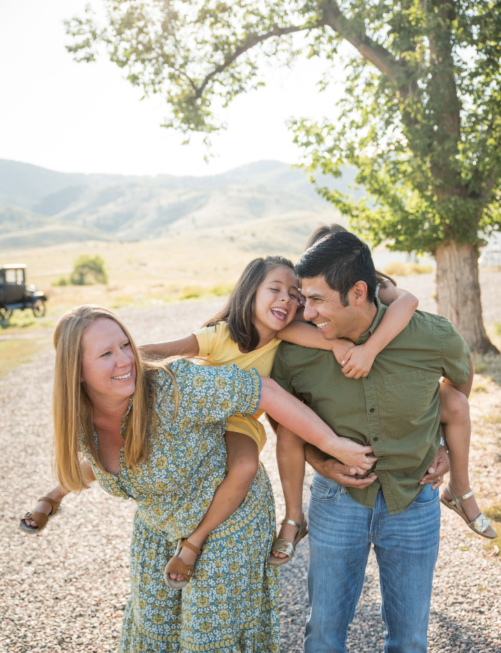 A mom and dad smile and laugh as they give their daughters piggyback rides, captured by Denver family photographer, Two One Photography.