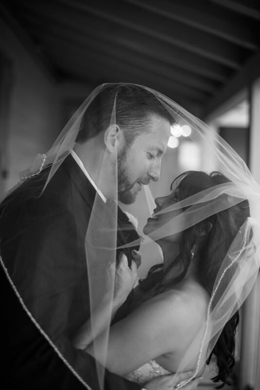 Black and white wedding photograph of bride and groom sharing an intimate moment under the bride's veil at the White Room in historic downtown St. Augustine. The couple is close together with the groom looking down at the bride, both smiling, with dramatic lighting and architectural elements visible in the background.