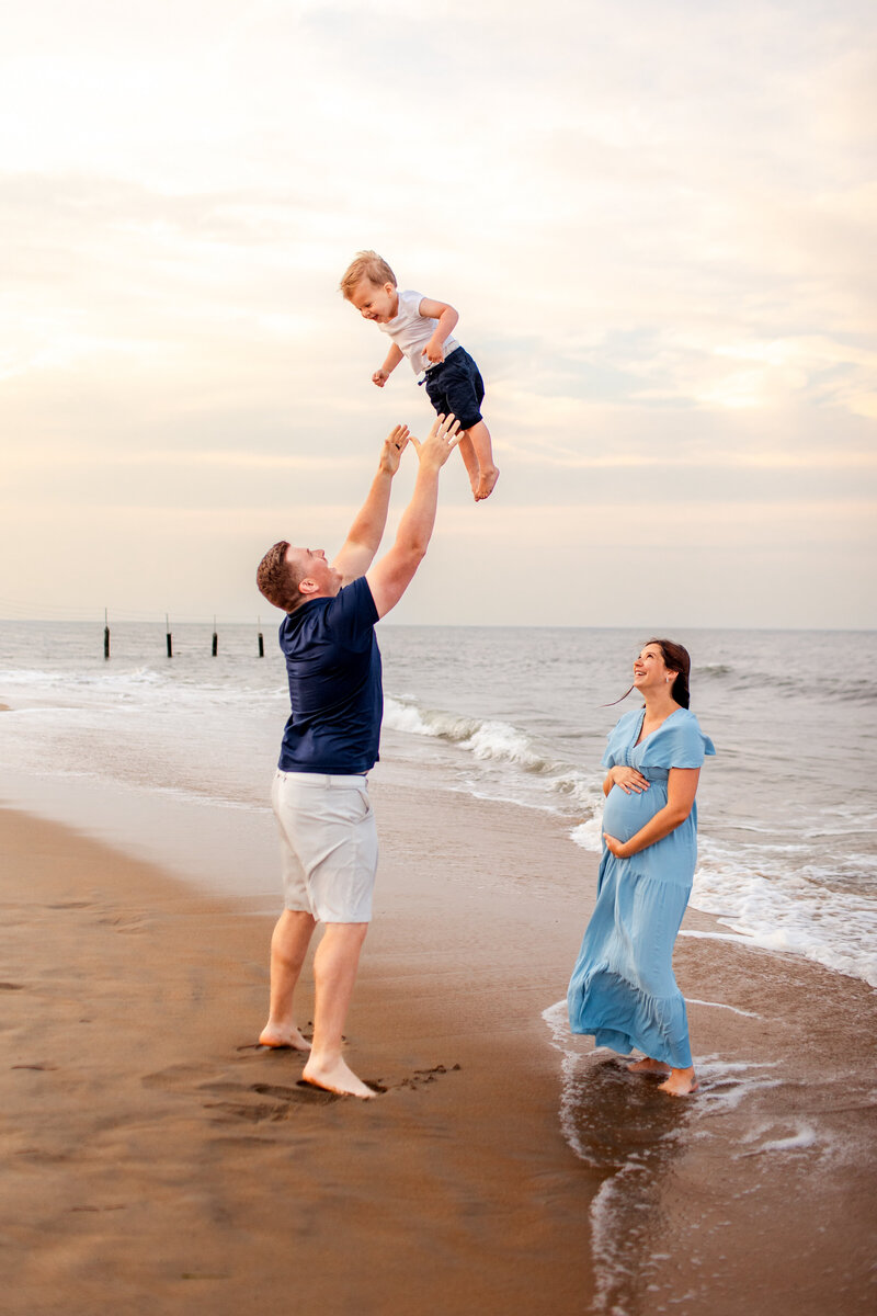 A young family of three plays at the water's edge during a joyful maternity session in Virginia Beach, as the mother cradles her belly and the father lifts their toddler into the air