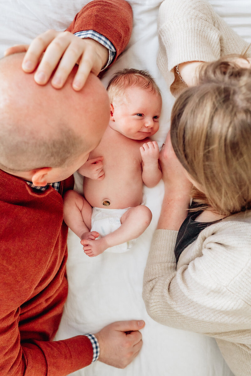A newborn baby lies between its parents on a bed, looking up at them with a small smile.