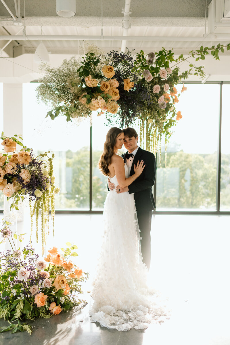 Bride and groom standing together beneath a dramatic hanging floral installation featuring garden-style roses, hydrangea, amaranthus, and greenery in soft peach, lavender, and cream tones. The couple embraces in a bright modern venue with floor-to-ceiling windows and natural light, showcasing an elegant, romantic wedding design with lush floral arrangements on the ground and overhead.