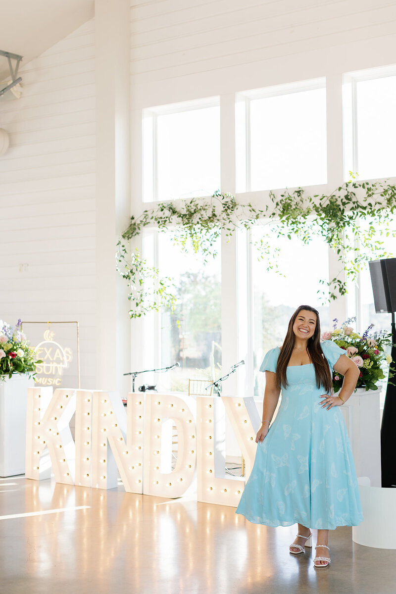 Makayla Rivera wearing a blue Show Me Your Mumu dress standing in front of wedding flowers and marquee light letters in a Fredericksburg Texas wedidng venue