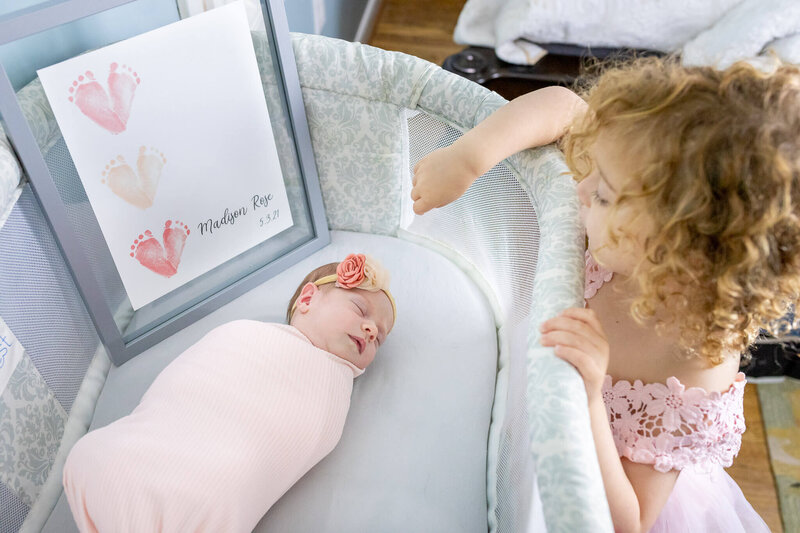 A girl peaking into a bassinet at her newborn sister during an Alexandria newborn photography session.