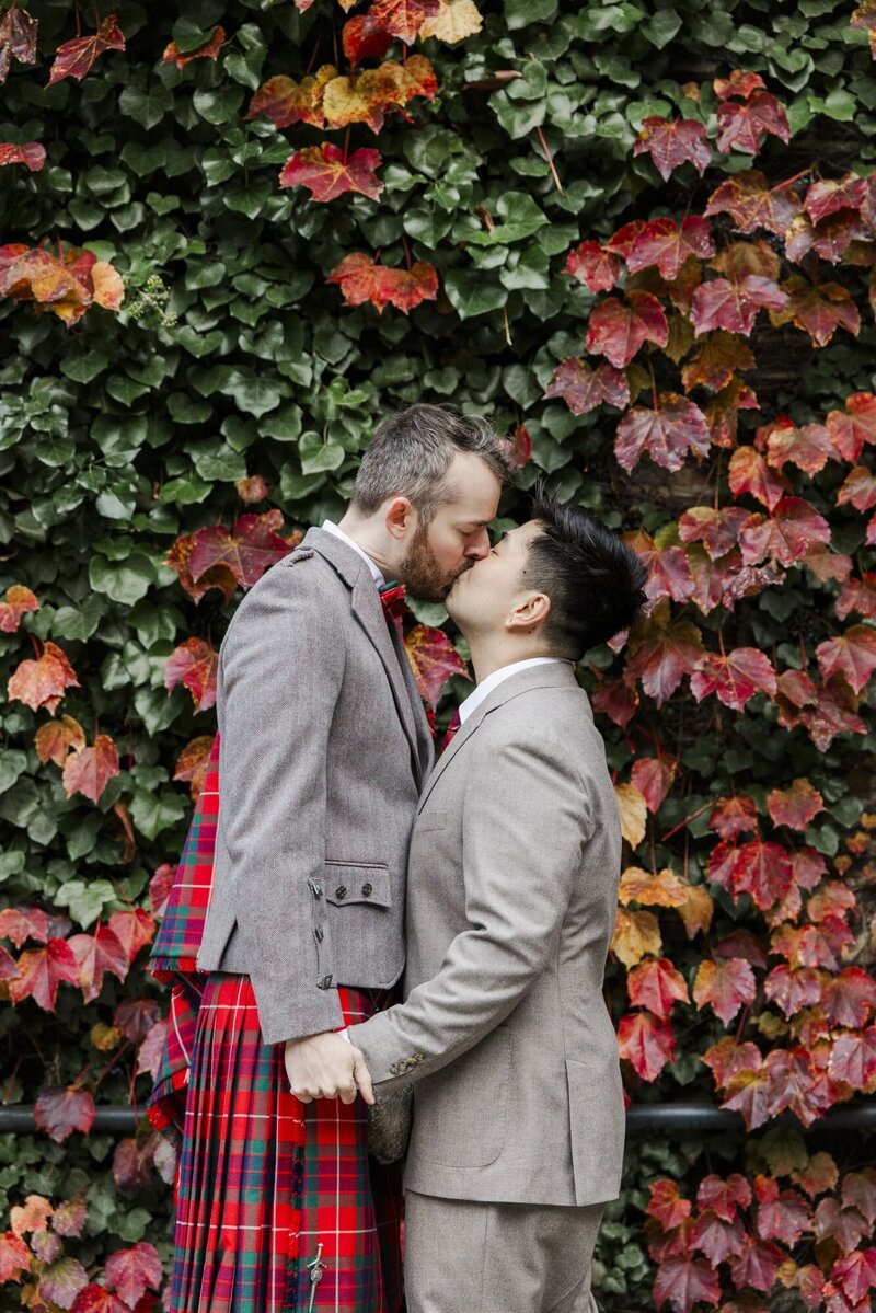 Two grooms kiss in front of a wall of autumn ivy at their Hudson Valley micro wedding