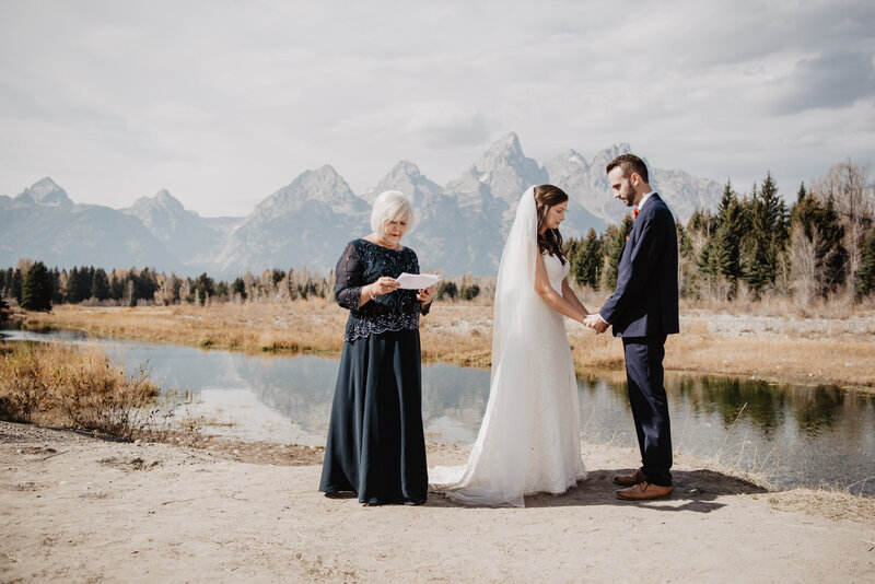Schwabacher Landing wedding location int he grand tetons with bride adn groom holding hands and praying with their wedding officiant on the bank of a lake with the mountains in the distance for their fall wedding