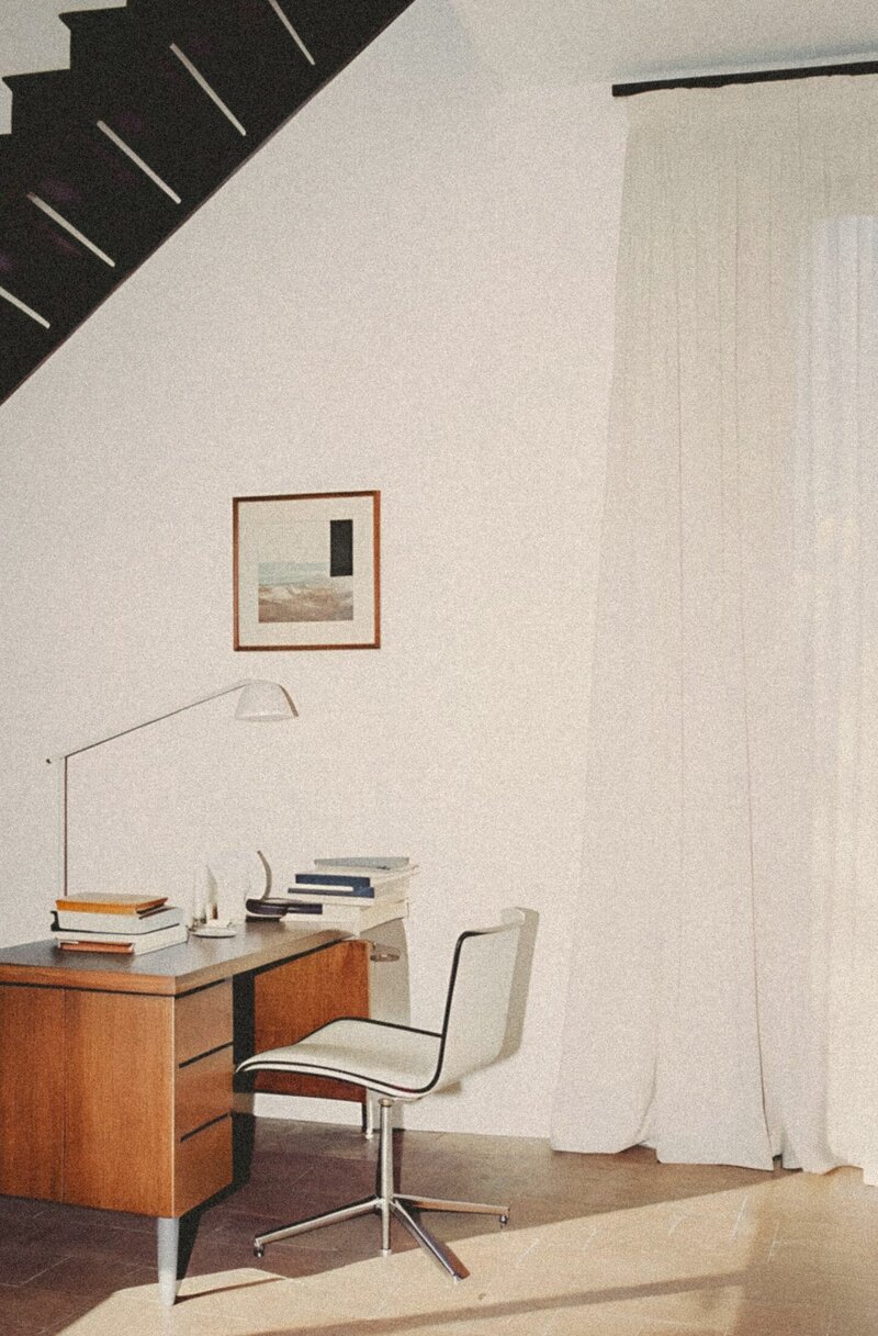 A minimalist white room featuring open black stairs, with a mid-century walnut desk and sleek steel chair beneath. Framed artwork and soft linen curtains add elegance to this thoughtfully curated home office space.
