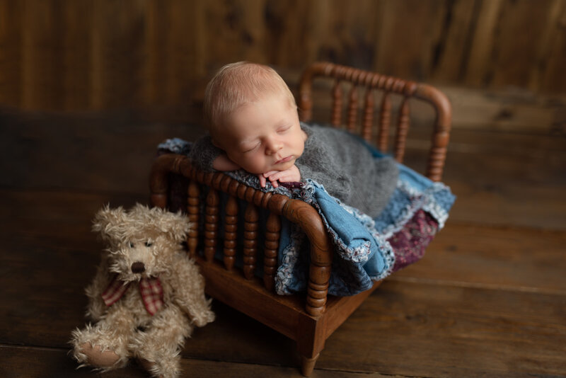 Newborn boy posed on a wooden spindle bed with vintage quilt and grey sleeper beside a plush bear – a rustic and heartfelt newborn portrait.