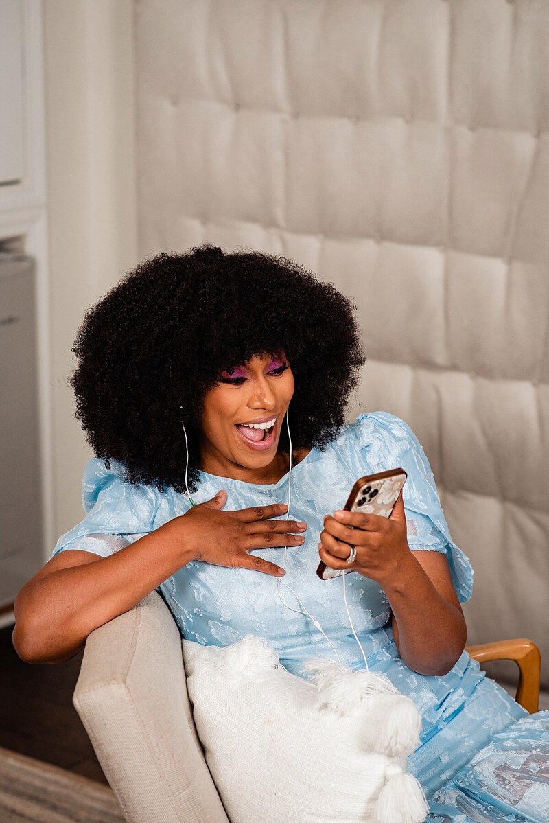 Sheena wearing a light blue dress, looking at her phone and laughing as she listens to a podcast
