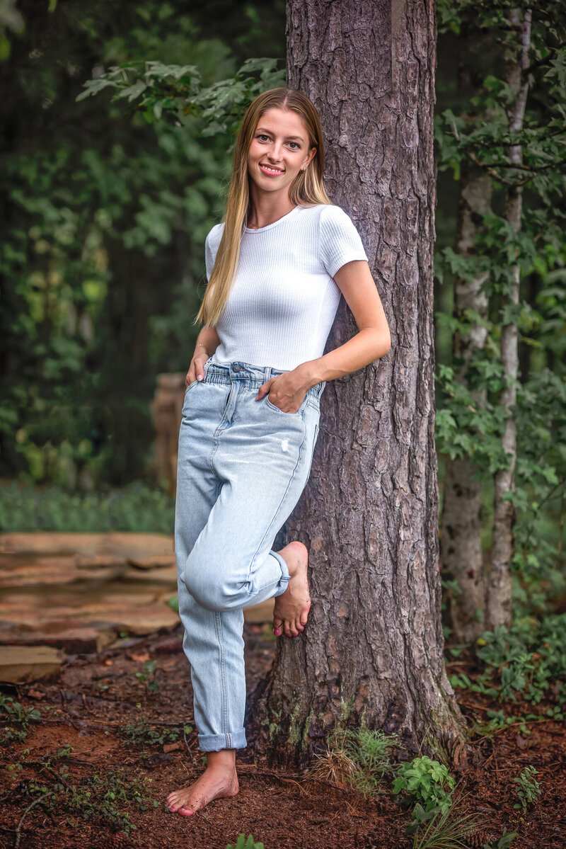 A teen girl with long blonde hair is leaning up against a tree and smiling for the camera.