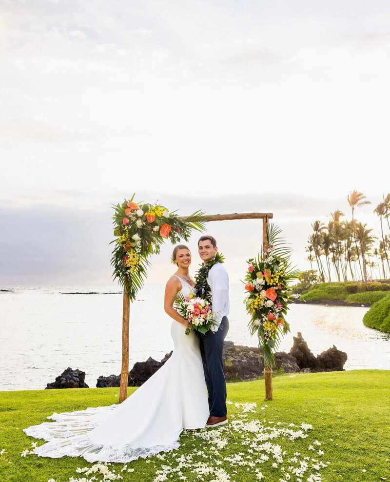 A couple standing under a tropical floral arch by the water