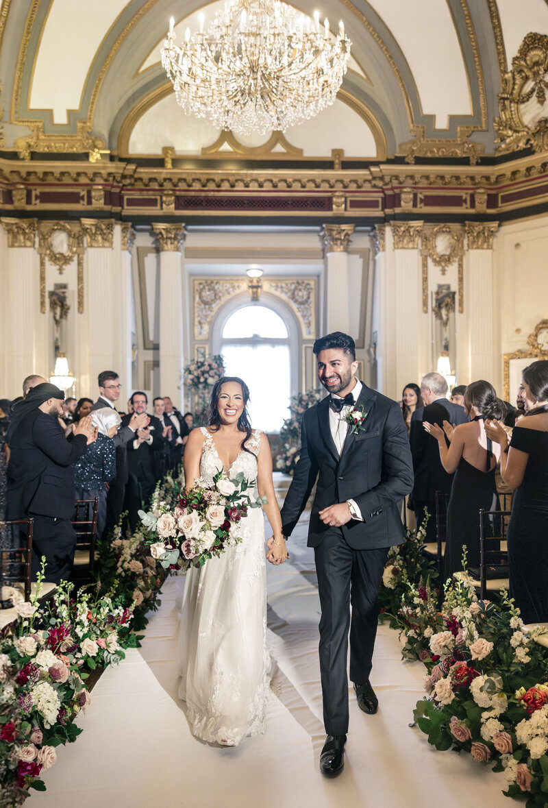 A bride and groom smile and hold hands while walking down the aisle in an ornate hall. She wears a white gown with a bouquet of flowers, and he wears a black tuxedo. Guests stand and clap on either side of the flower-lined aisle under a grand chandelier.