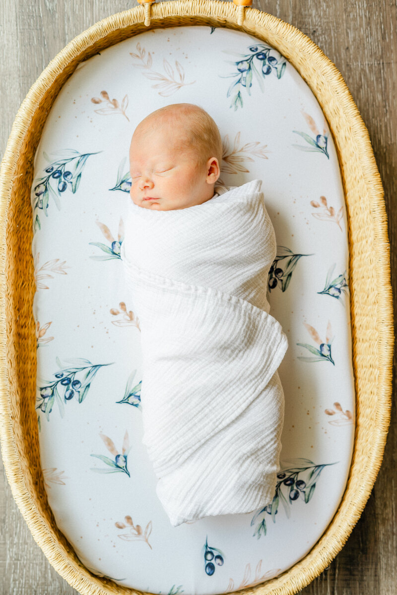a newborn swaddled in a white blanket sleeps in a Moses basket on top of a floral mattress sheet.